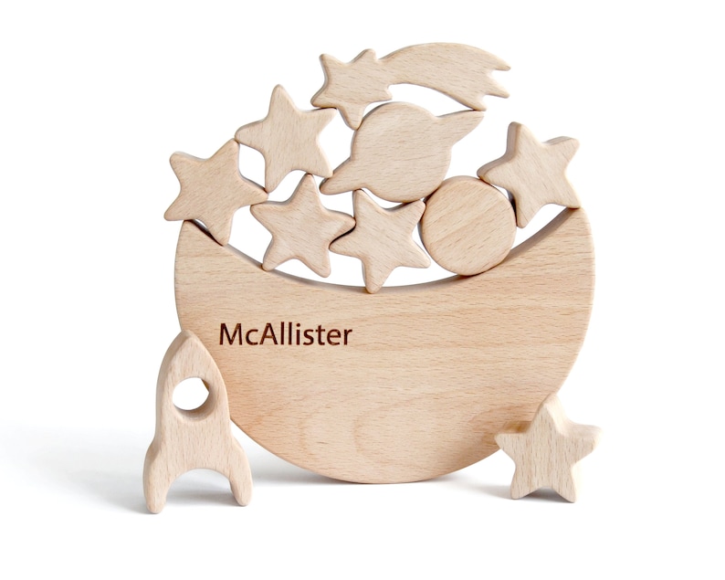 Wooden moon stacking and balance game in natural beech wood, crescent moon rocker holding six stars, a shooting star comet, planet and rocket, unpainted Waldorf style fine motor toy for toddlers, space theme nursery shelf decor, white background.