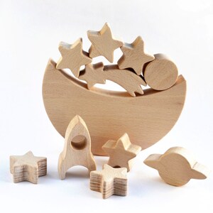 Handmade unpainted beechwood moon balance toy with stars, planets and a rocket balanced on a crescent moon base, open ended wooden stacking toy for toddlers, eco friendly first birthday gift and moon nursery decor, plain white background.