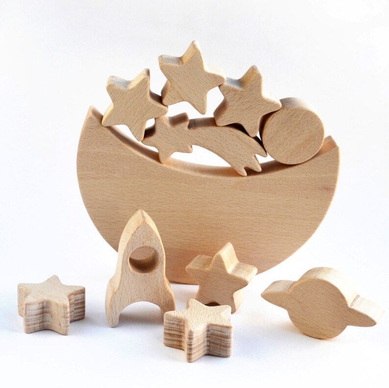 Handmade unpainted beechwood moon balance toy with stars, planets and a rocket balanced on a crescent moon base, open ended wooden stacking toy for toddlers, eco friendly first birthday gift and moon nursery decor, plain white background.