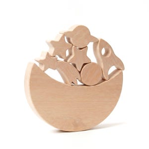 Pu&ograve; includere: Un set di blocchi impilabili in legno chiaro con forme a tema spaziale, tra cui una base a mezzaluna, un razzo, un pianeta con anelli, un disco circolare, due stelle e una stella cadente, disposti in una pila verticale bilanciata.