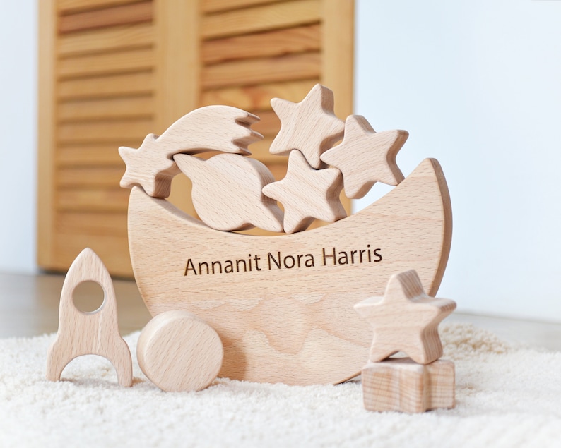 Pu&ograve; includere: Un set di giocattoli impilabili in legno chiaro con una base a forma di mezzaluna incisa con il nome Annanit Nora Harris, accompagnato da varie forme a tema spaziale tra cui stelle, un pianeta con anelli, un razzo e un disco circolare.