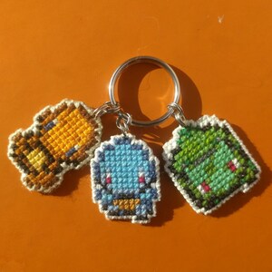 May include: A silver key chain with three cross-stitched charms. The charms are a brown and orange character, a blue character, and a green character.