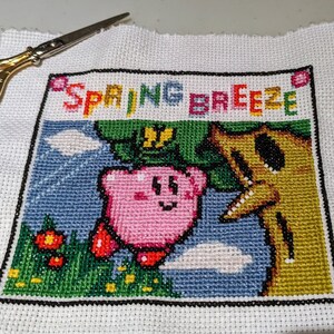 May include: Cross-stitch embroidery of a pink character with a yellow flower and a brown tree, with the text "SPRING BREEZE" above.