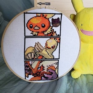 May include: A white embroidery hoop with a cross-stitch design of three Pokemon characters. The characters are a fire-type Pokemon, a bird-type Pokemon, and a cat-type Pokemon. A yellow plush toy of a Pokemon character is in the background.