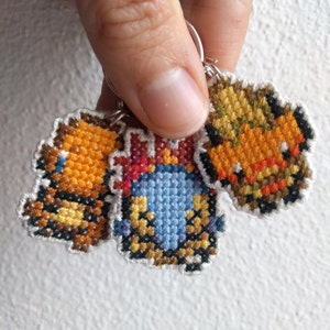 May include: Three small cross-stitched keychains featuring cartoon characters. The keychain on the left is brown and orange, the middle keychain is blue, red, and yellow, and the keychain on the right is orange and brown.