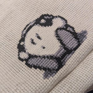 May include: A close-up of a cross-stitch project showing a cartoon character in grey and white. The character is stitched on cream-coloured fabric. White thread is visible at the top, indicating the embroidery is in progress.
