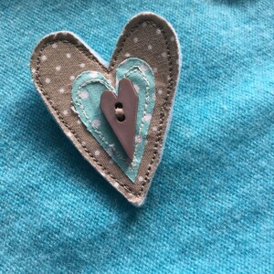 May include: A brown heart-shaped fabric patch with a smaller blue heart-shaped fabric patch inside. The larger patch has white polka dots and the smaller patch has white polka dots. A brown button is sewn in the center of the smaller patch.