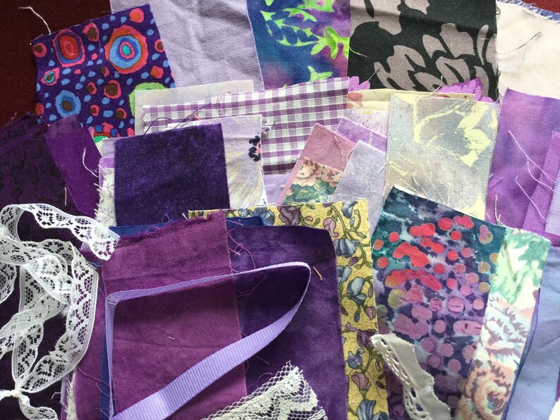 May include: A collection of purple and lavender fabric scraps, including floral prints, gingham, and solid colors. There is also white lace and a purple ribbon.