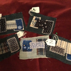 May include: Four denim squares with different fabric patches sewn on top. Each square has a white tag with a letter on it: V, X, N, and Z.