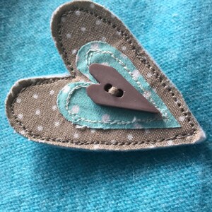 May include: A brown heart-shaped fabric patch with white polka dots, sewn onto a turquoise fabric background. A smaller turquoise heart with white polka dots is sewn inside the larger heart, and a brown heart-shaped button is sewn in the center.