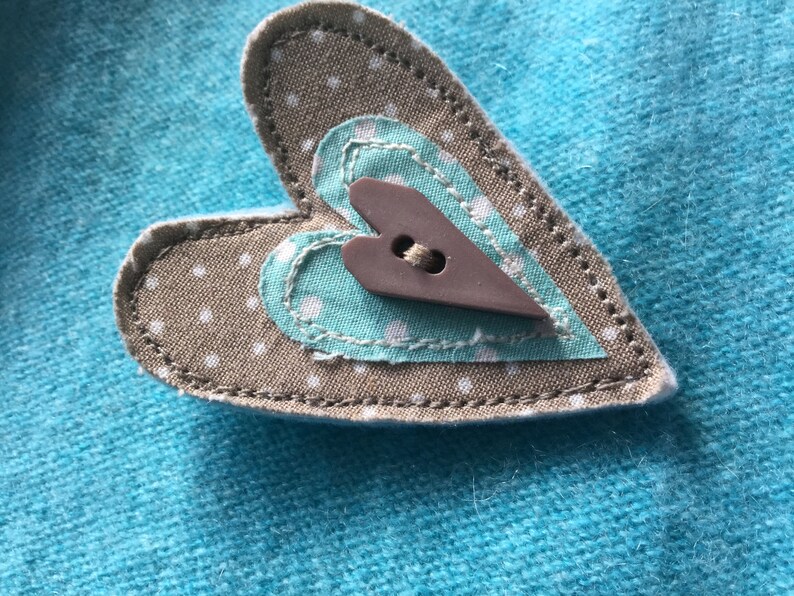 May include: A brown heart-shaped fabric patch with white polka dots, sewn onto a turquoise fabric background. A smaller turquoise heart with white polka dots is sewn inside the larger heart, and a brown heart-shaped button is sewn in the center.