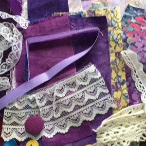 May include: A collection of purple fabric scraps, lace trim, and ribbon. The fabric scraps are in various shades of purple, and the lace trim is white. There are also two purple buttons and a yellow button.