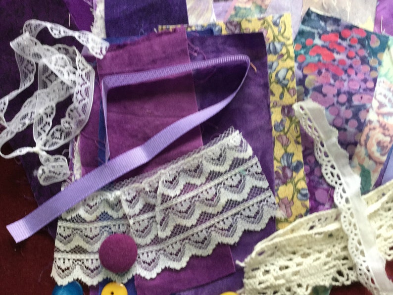 May include: A collection of purple fabric scraps, lace trim, and ribbon. The fabric scraps are in various shades of purple, and the lace trim is white. There are also two purple buttons and a yellow button.