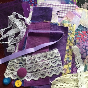May include: A collection of purple fabric scraps, lace trim, and buttons. The fabric scraps are in various shades of purple, including lavender, lilac, and amethyst. There is also a spool of purple thread and a piece of white lace.