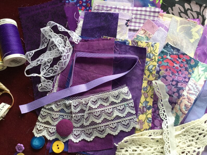 May include: A collection of purple fabric scraps, lace trim, and buttons. The fabric scraps are in various shades of purple, including lavender, lilac, and amethyst. There is also a spool of purple thread and a piece of white lace.