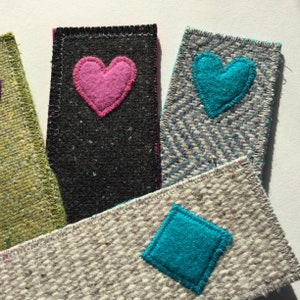 Puede incluir: Cuatro marcapáginas de tela con diferentes patrones y colores. Un marcapáginas es negro con un corazón rosa, otro es azul con un corazón turquesa, otro es verde con un corazón blanco y el último es blanco con un rombo turquesa.