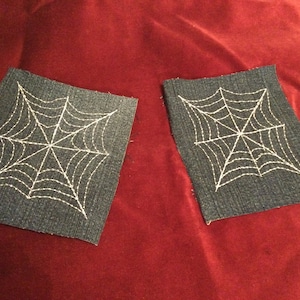 May include: Two denim patches with white embroidered spiderwebs. The patches are on a red velvet background.