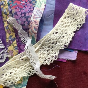 May include: A collection of purple fabric scraps and white lace trim. The fabric scraps are in various shades of purple and have a textured, hand-dyed look. The lace trim is delicate and intricate.