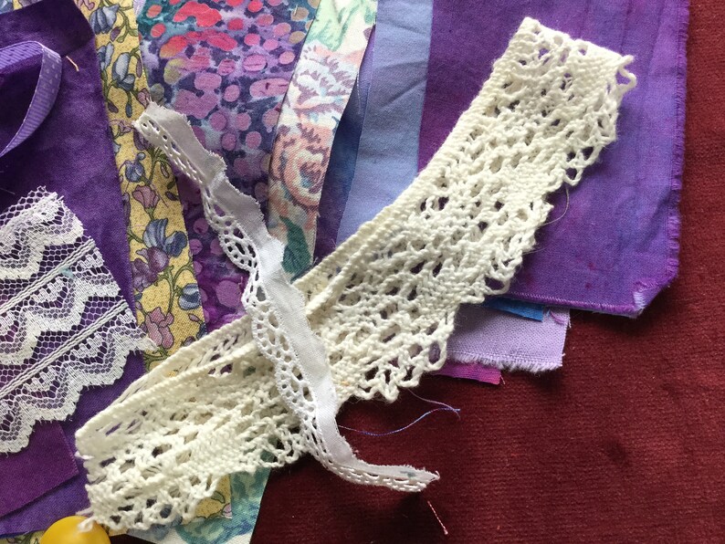May include: A collection of purple fabric scraps and white lace trim. The fabric scraps are in various shades of purple and have a textured, hand-dyed look. The lace trim is delicate and intricate.