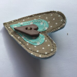 May include: A brown heart-shaped fabric pin with a brown button in the center. The fabric is beige with white polka dots and has a turquoise heart sewn on top.