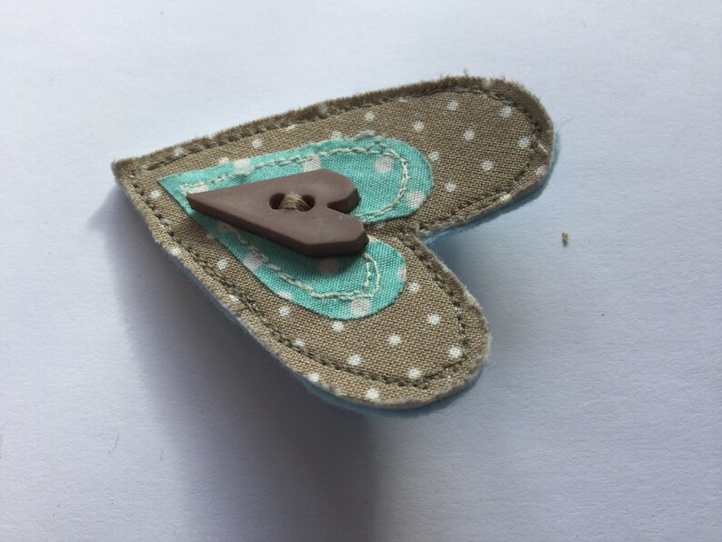 May include: A brown heart-shaped fabric pin with a brown button in the center. The fabric is beige with white polka dots and has a turquoise heart sewn on top.