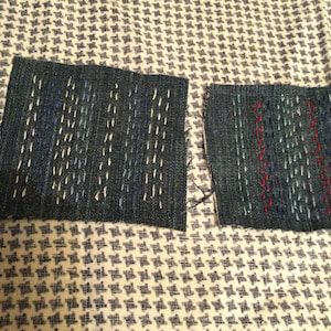 May include: Two dark denim fabric squares with white, blue, and red stitching. The squares are on a white and blue patterned fabric. The stitching creates a textured effect, suggesting a craft or repair project.