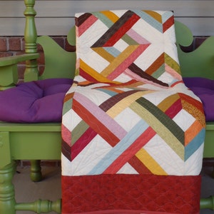 May include: A colourful patchwork quilt with a red, white and green pattern is draped over a green bench with a purple cushion. The quilt has a red border and is made of various fabrics.