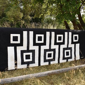 May include: Black and white quilt with a geometric pattern of squares and rectangles. The quilt is hanging on a wooden fence.