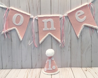 May include: Pink felt banner with white letters spelling "one" and white pearl trim. A pink felt party hat with white polka dots and a white felt "1" is in the foreground.