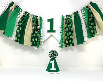 May include: Green and gold first birthday party decorations with a banner that says "1" and a green party hat with a white pom-pom and the number "1" on it.