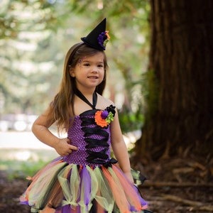 K&ouml;nnte beinhalten: Ein Kind in einem Halloween-Hexenkost&uuml;m. Das Outfit umfasst ein lila-schwarzes Oberteil, einen bunten Tutu-Rock und einen schwarzen Hexenhut. Das Kind h&auml;lt einen schwarzen Kessel mit Sternaussparungen.