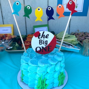May include: A blue and white cake decorated with a red and white circle that says "the Big ONE". The cake is surrounded by green frosting that looks like seaweed. There are also colorful felt fish hanging from a string above the cake.