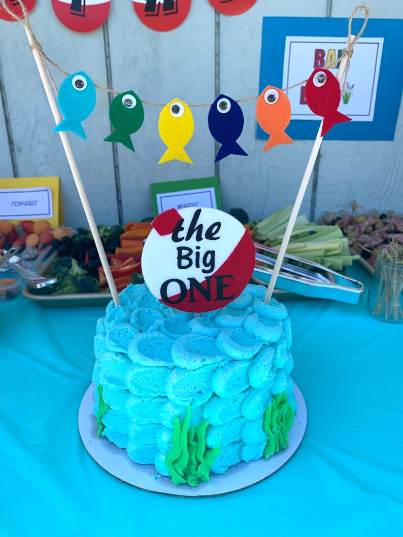 May include: A blue and white cake decorated with a red and white circle that says "the Big ONE". The cake is surrounded by green frosting that looks like seaweed. There are also colorful felt fish hanging from a string above the cake.