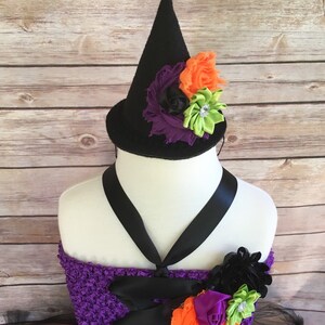 K&ouml;nnte beinhalten: Ein Halloween-Kost&uuml;m mit einem schwarzen Hexenhut, der mit orangefarbenen, lila und gr&uuml;nen Stoffblumen verziert ist. Das Kost&uuml;m umfasst ein lilafarbenes H&auml;keloberteil mit schwarzen Schleifenakzenten und einen schwarzen T&uuml;llrock. Das Gesamtthema ist ein festliches Halloween-Outfit.