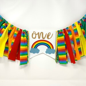 May include: A colorful banner with the word "ONE" in gold glitter lettering, a rainbow, and blue clouds. The banner is decorated with rainbow-colored fabric strips and is perfect for a first birthday celebration.
