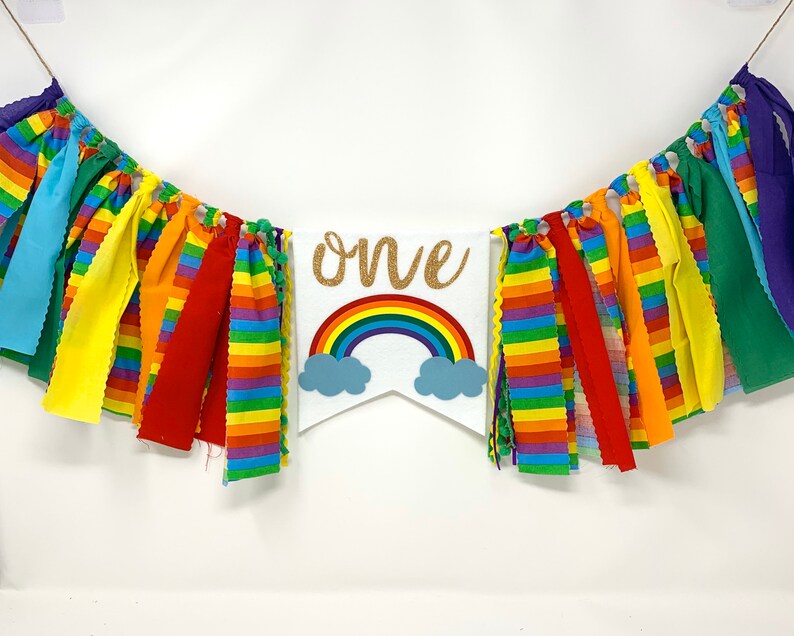 May include: A colorful banner with the word "ONE" in gold glitter lettering, a rainbow, and blue clouds. The banner is decorated with rainbow-colored fabric strips and is perfect for a first birthday celebration.