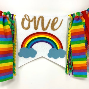 May include: A white banner with the word "one" in gold glitter script, a rainbow, and blue cloud accents. The banner is decorated with colorful striped fabric and green pom-poms. A festive decoration for a birthday celebration.