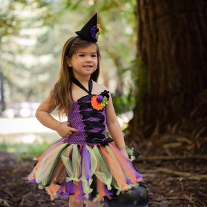 K&ouml;nnte beinhalten: Ein Halloween-Hexenkost&uuml;m f&uuml;r Kinder. Das lila Oberteil ist mit schwarzem Band und einem passenden Tutu-Rock verziert. Das Outfit beinhaltet einen schwarzen Hexenhut mit floralen Akzenten und einen schwarzen Kessel.