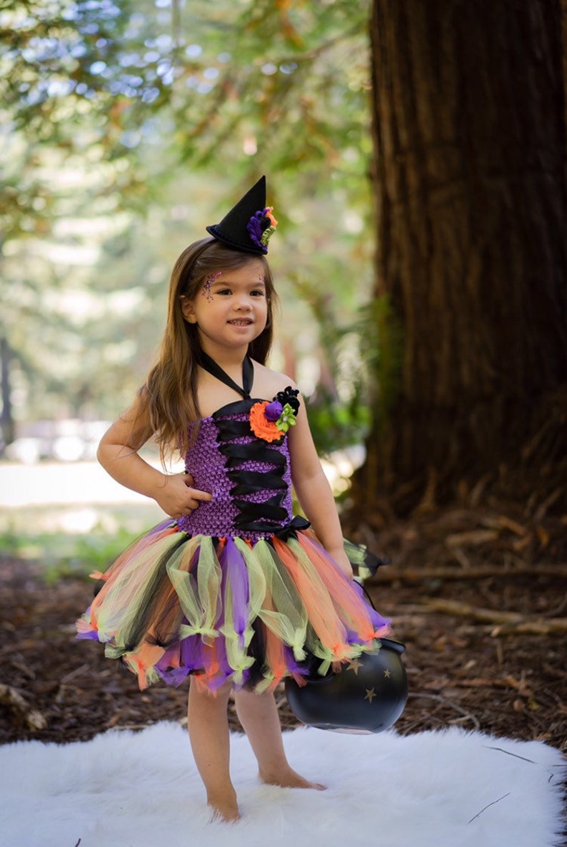 K&ouml;nnte beinhalten: Ein Halloween-Hexenkost&uuml;m f&uuml;r Kinder. Das lila Oberteil ist mit schwarzem Band und einem passenden Tutu-Rock verziert. Das Outfit beinhaltet einen schwarzen Hexenhut mit floralen Akzenten und einen schwarzen Kessel.