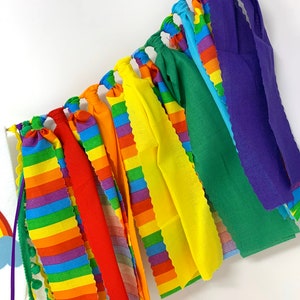 May include: A colorful rainbow-themed garland with fabric strips in various colors, including red, orange, yellow, green, blue, and purple. The garland is suspended from a string and is suitable for party decorations or festive occasions.
