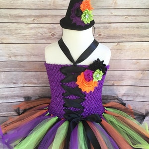 K&ouml;nnte beinhalten: Ein lila geh&auml;keltes Oberteil-Tutu-Kleid mit einem schwarzen Korsett-Design. Der Rock hat Lagen aus orangefarbenem, gr&uuml;nem, lila und schwarzem T&uuml;ll. Ein passender schwarzer Hexenhut mit orangefarbenen, lila und gr&uuml;nen floralen Akzenten vervollst&auml;ndigt das Halloween-Kost&uuml;m.