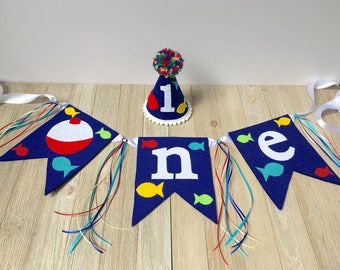May include: A blue felt banner with the word "one" spelled out in white felt letters. The banner is decorated with red, yellow, and blue fish and a red and white fishing bobber. The banner is strung with white ribbon and colorful yarn. A blue felt party hat with a white felt number "1" and a colorful pom-pom on top is also included.