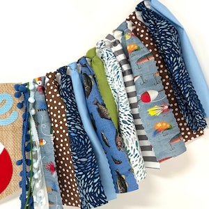 May include: A blue and brown fabric banner with a fishing theme. The banner has various patterns including fish, fishing lures, and polka dots. The banner is strung together with white yarn.