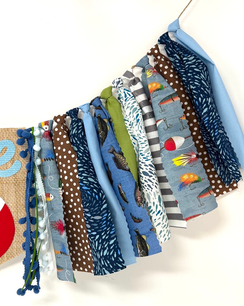 May include: A blue and brown fabric banner with a fishing theme. The banner has various patterns including fish, fishing lures, and polka dots. The banner is strung together with white yarn.