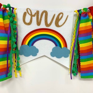 May include: A white banner with the word "one" in gold glitter, a rainbow, and blue cloud cutouts. The banner is decorated with colorful ribbons and green pom-poms, creating a festive look. Perfect for a first birthday celebration.