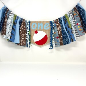 May include: A blue and brown fabric banner with the word "one" and a red and white fishing bobber. The banner is decorated with white pom-poms.