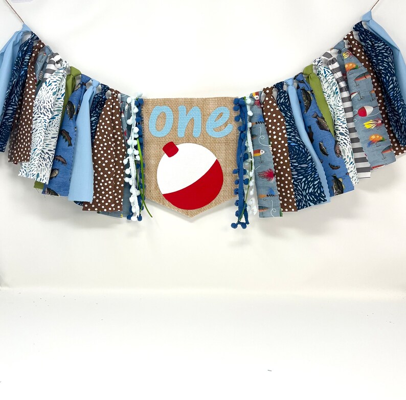 May include: A blue and brown fabric banner with the word "one" and a red and white fishing bobber. The banner is decorated with white pom-poms.