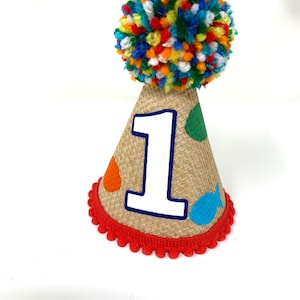 May include: A brown burlap party hat with a red pom-pom on top. The hat has a white number "1" on the front and colorful fish and dots.