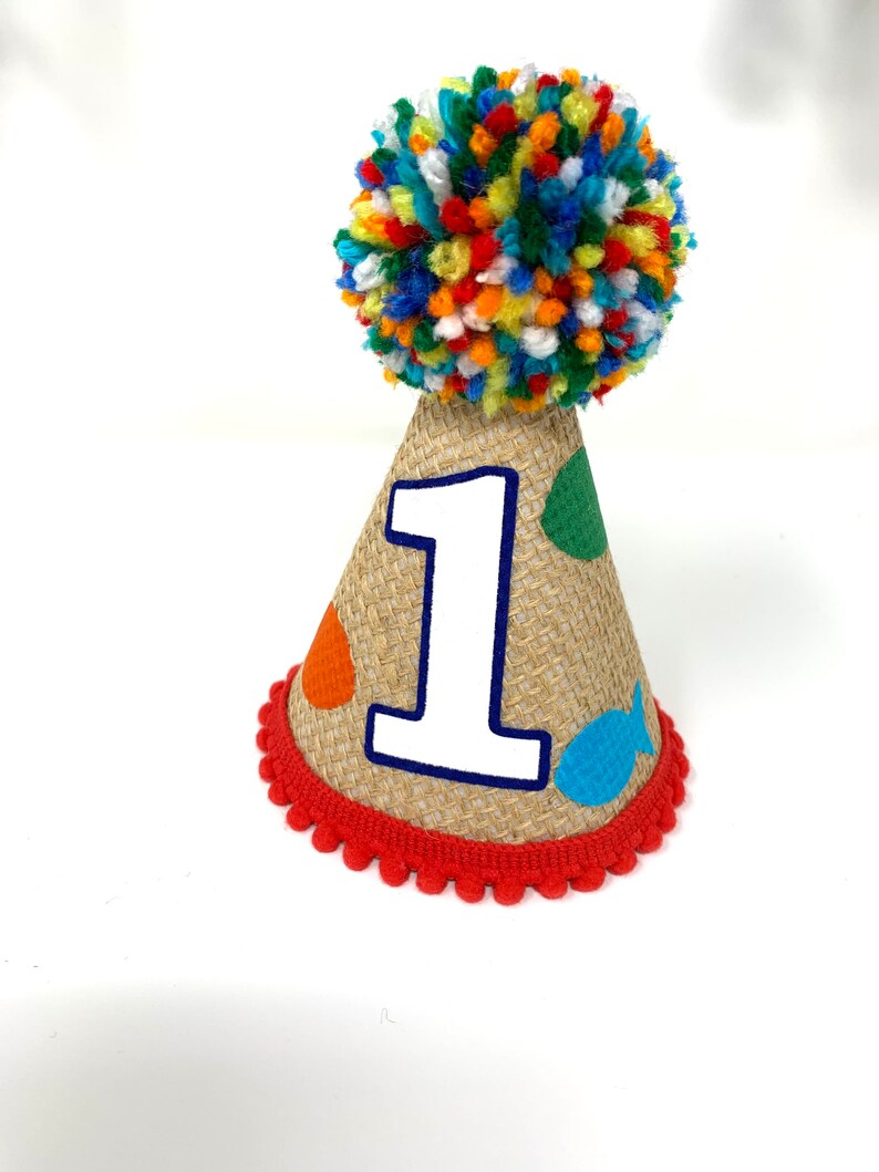 May include: A brown burlap party hat with a red pom-pom on top. The hat has a white number "1" on the front and colorful fish and dots.