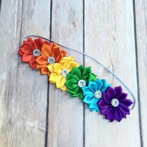 Puede incluir: Diadema arco&iacute;ris con seis flores de sat&eacute;n, cada una con un centro de pedrer&iacute;a. Las flores son rojas, naranjas, amarillas, verdes, azules y moradas.